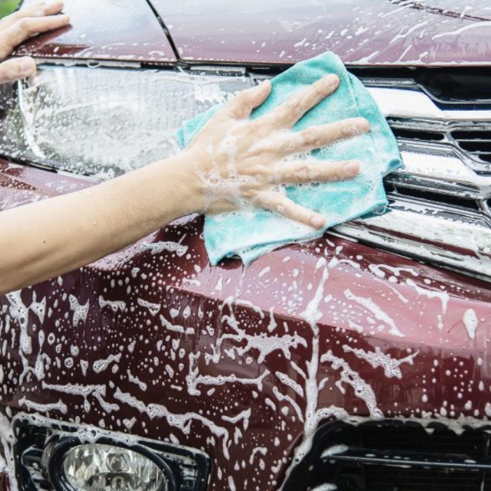 Close-up of a person's soapy hand using a turquoise microfiber cloth to wipe the front hood and grille area of a maroon car. The car surface is covered in white soap suds, which are dripping down.