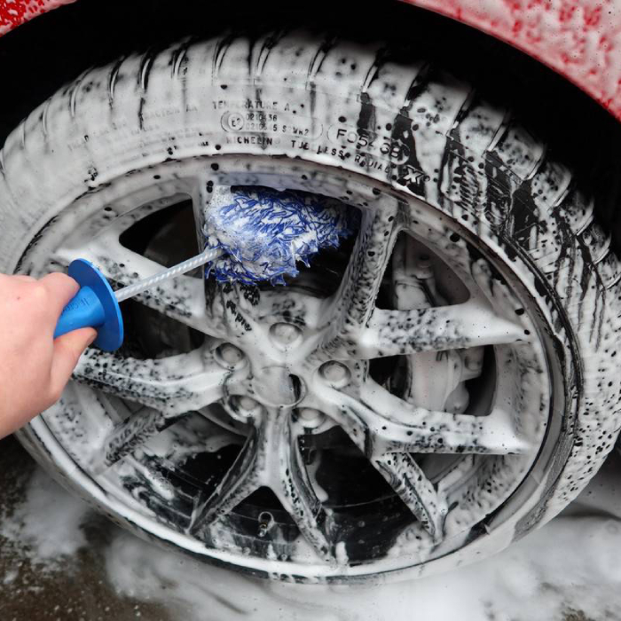 Close-up of a person's hand using a blue-handled, blue and white microfiber wheel brush to scrub the multi-spoke alloy wheel of a car. Both the wheel and tire are covered in thick white soap suds.
