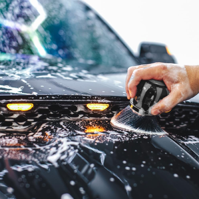 Close-up of a soapy hand holding a round, black detailing brush with soft bristles, cleaning the suds-covered hood and grille area of a black vehicle. The grille features illuminated amber marker lights.
